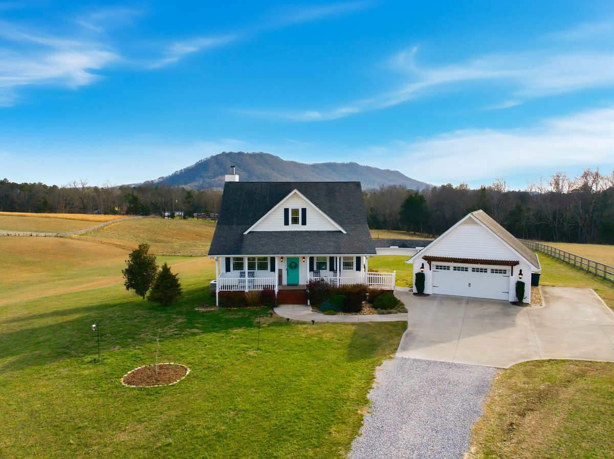 Aerial view of a farmhouse with a dark roof and a white garage on open pasture with a mountain peak behind, Corryton TN