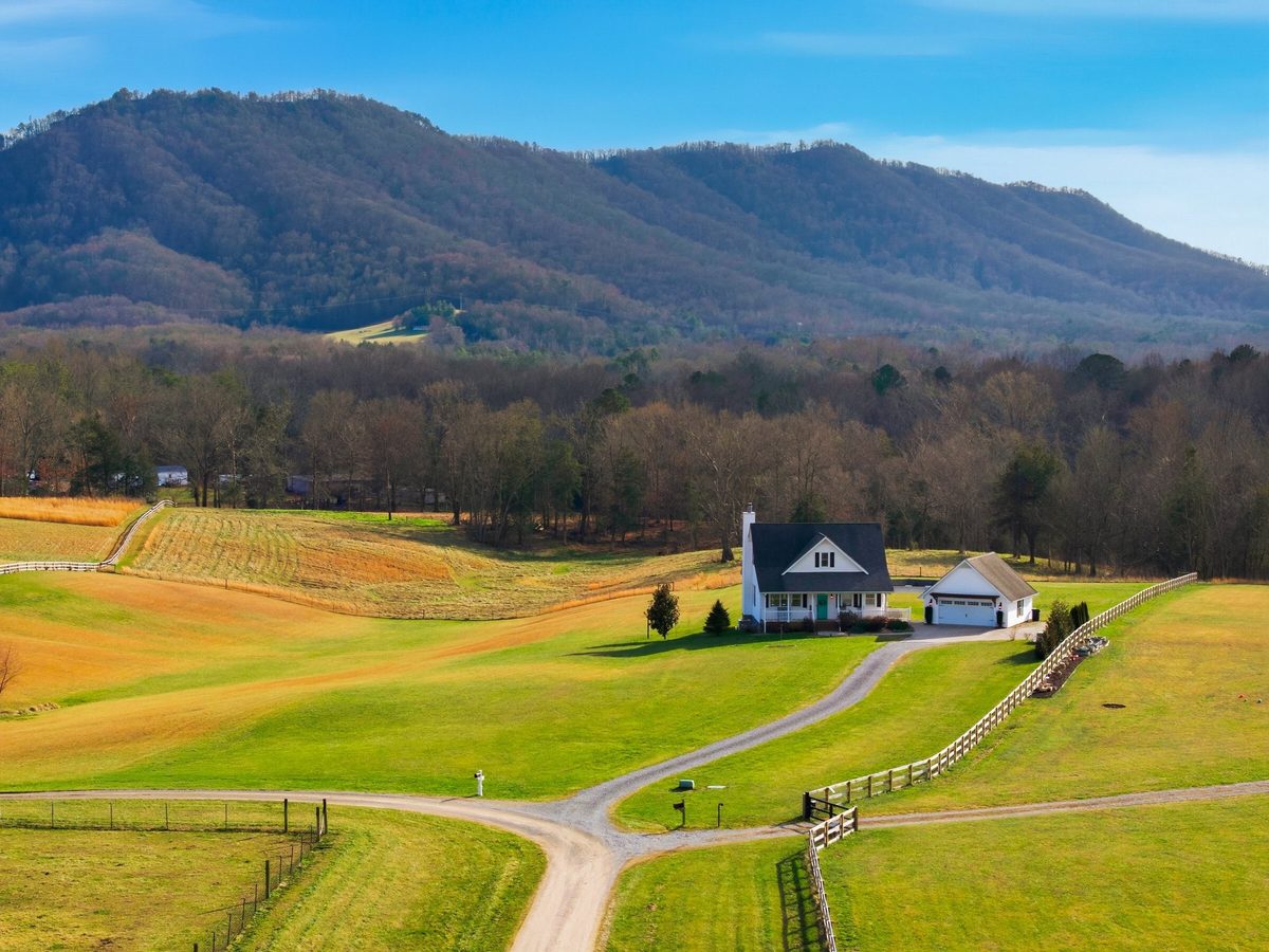 Aerial view of a white farmhouse and garage among green fields with a gravel drive, fenced pasture and a mountain ridge behind, Corryton TN