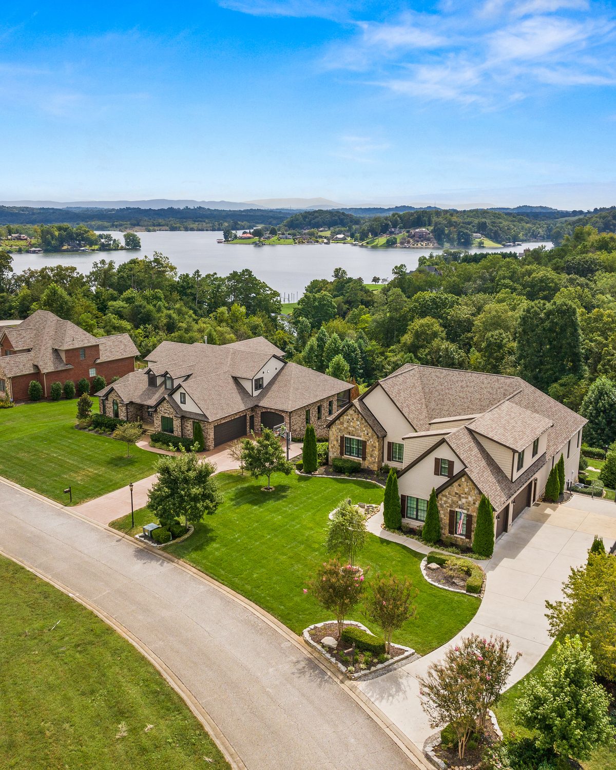 Aerial view of two stone and siding homes on a curving street with green lawns, a lake and wooded ridges behind, Knoxville TN